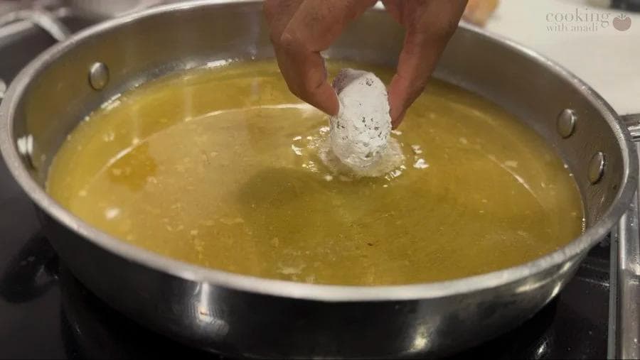Hand placing egg-coated potato fritter into hot oil for frying in a stainless steel pan.