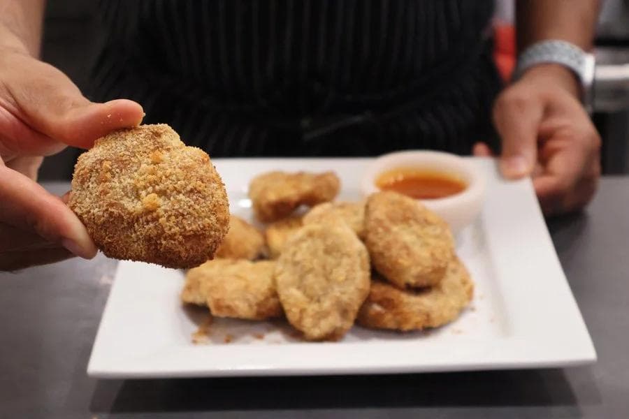 Hand holding a golden crunchy air fryer chicken nugget above a white plate with more nuggets and dipping sauce.