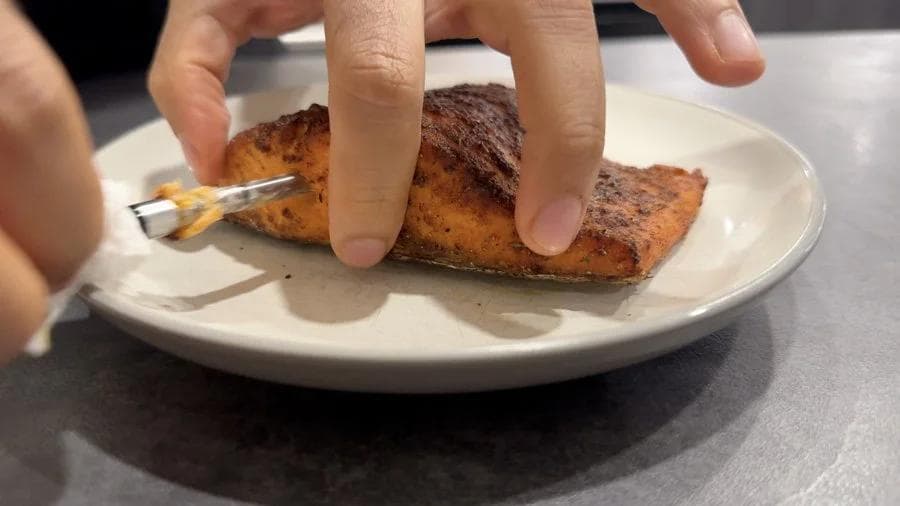 Hands peeling skin off cooked salmon fillet on white plate with fork.