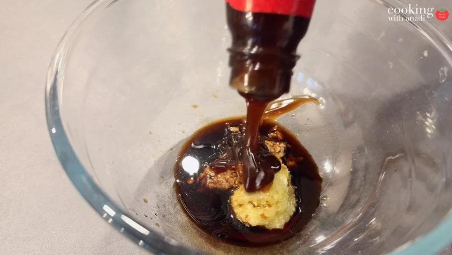Dark soy sauce being poured over garlic paste and other marinade ingredients in a glass bowl.