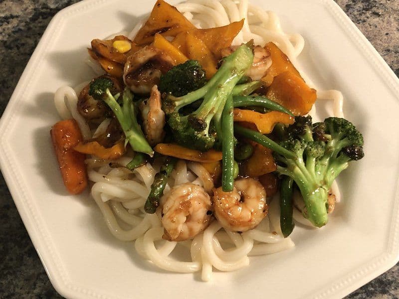 Teriyaki shrimp stir fry served over udon noodles with broccoli, mushrooms, and bell peppers on a white plate.