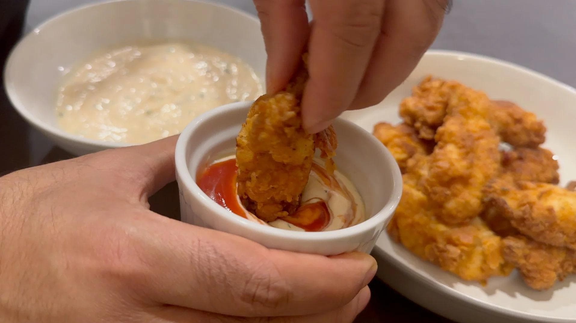 Crispy fried chicken tender being dipped into ketchup with creamy dipping sauce and more tenders on plate nearby.