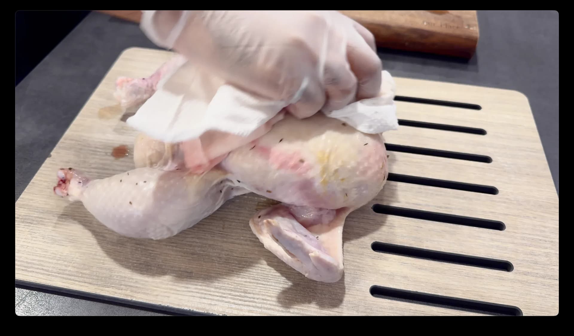 Hands in gloves making slits in raw chicken legs on a wooden cutting board with black grooves.