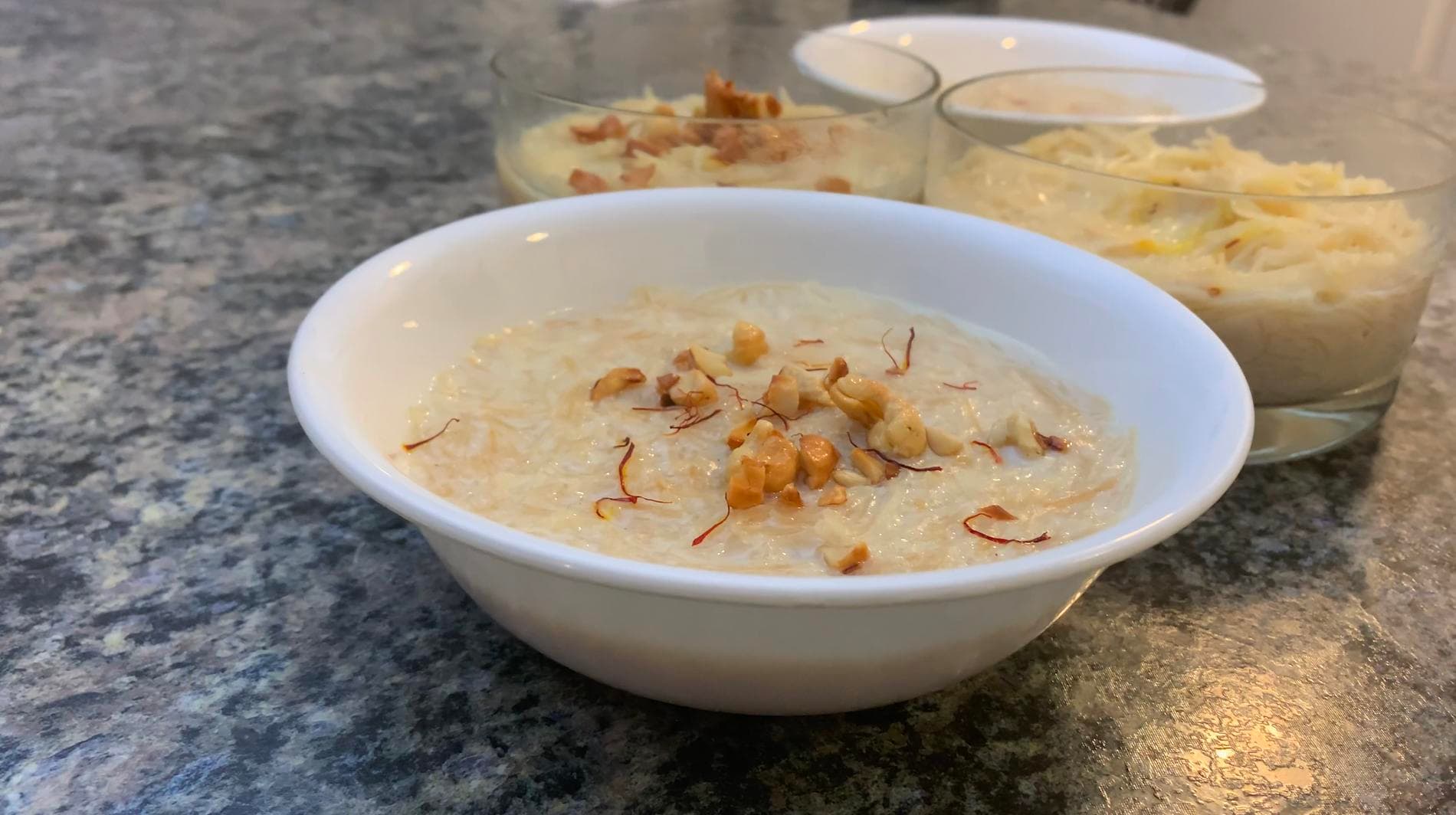 Seviyan kheer served in a white bowl, topped with chopped nuts and saffron strands, with additional servings in glasses behind.