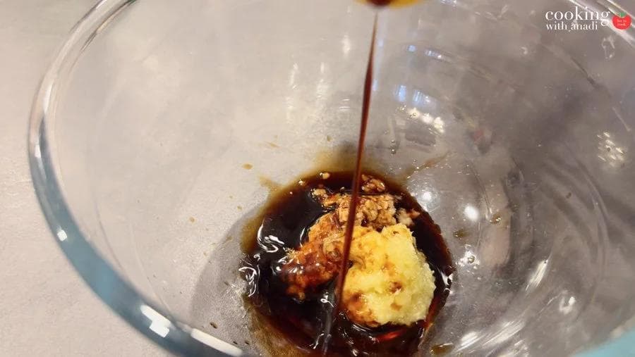 Dark soy sauce being poured over garlic-lemongrass paste mixture in a clear glass bowl.