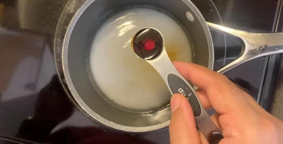 Hand holding a measuring spoon with vanilla extract over a saucepan of dissolved sugar and water mixture.