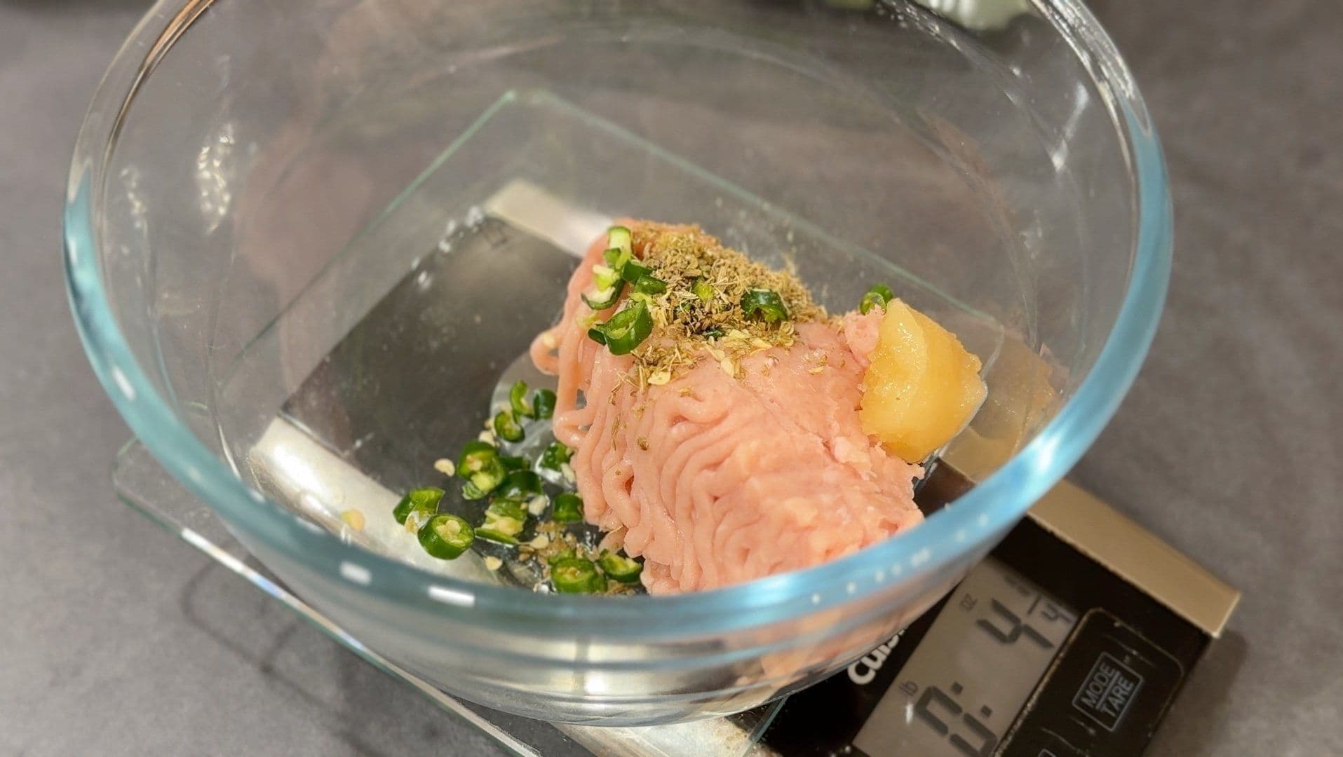 Ground meat mixture with diced green peppers, pineapple, and spices in glass bowl on kitchen scale