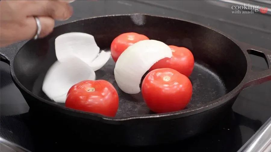 Whole red tomatoes and white onion halves charring in a black cast iron skillet on stovetop.
