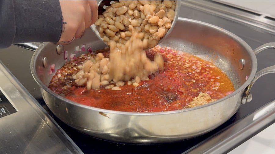 Cooked beans being poured into a simmering red tomato sauce in a metal pan on the stovetop.