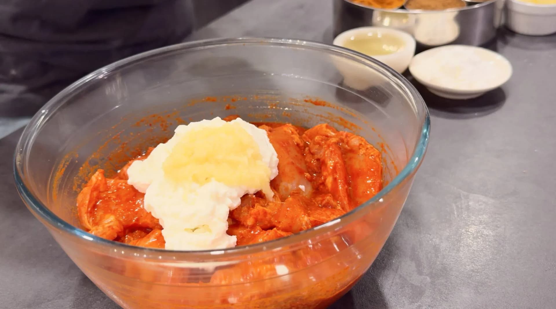Glass bowl of red-marinated chicken pieces topped with yogurt and butter, ready to be mixed for second marinade.
