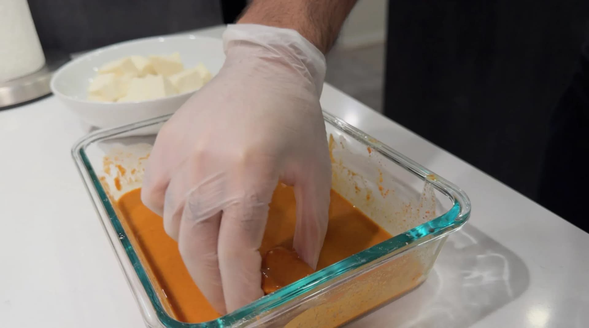 Gloved hand pressing paneer cubes into orange chilli batter in a glass baking dish, with cubed paneer in a bowl behind.