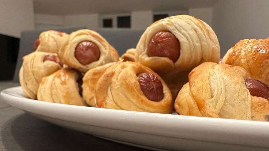 Air fryer pigs in a blanket arranged on a white plate, showing golden crescent roll wrapping around cocktail sausages.