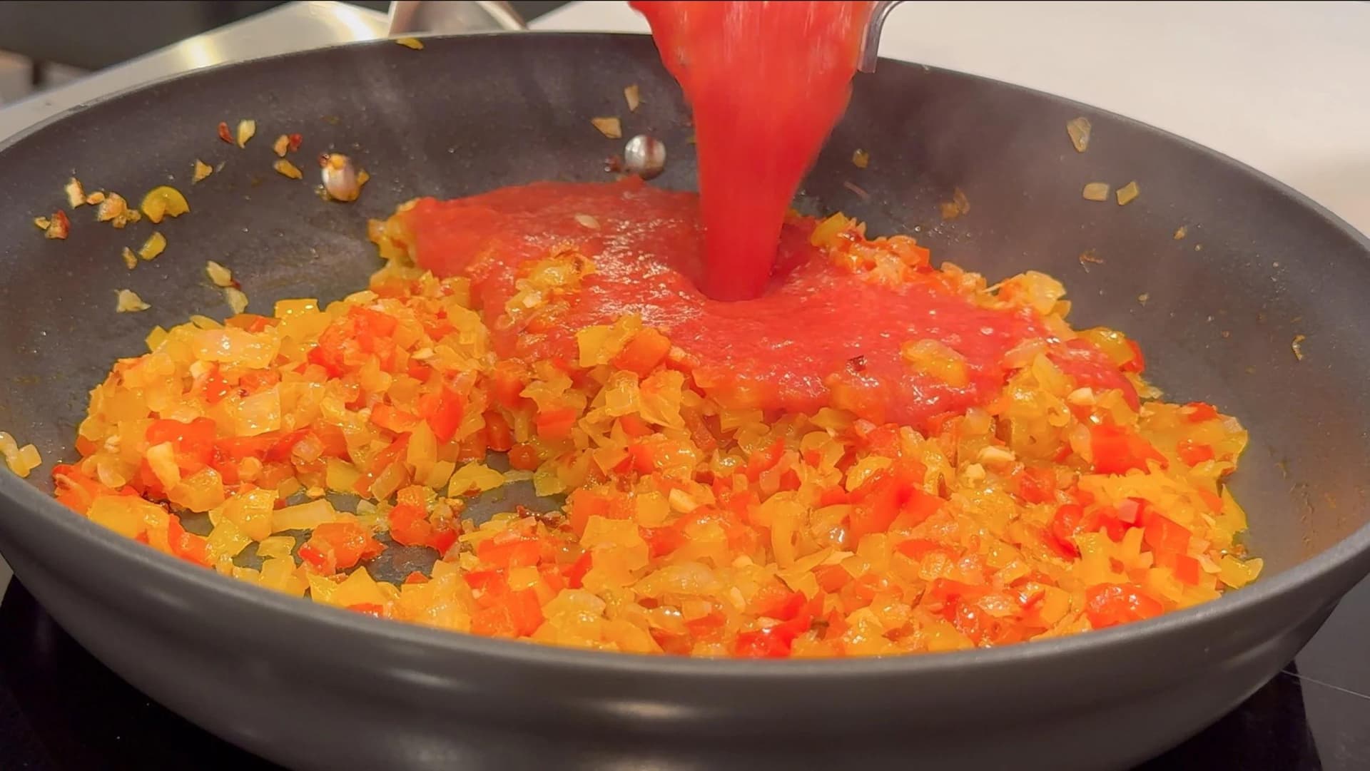 Crushed tomatoes being poured from a red bowl into a skillet of sautéed diced bell peppers and onions.