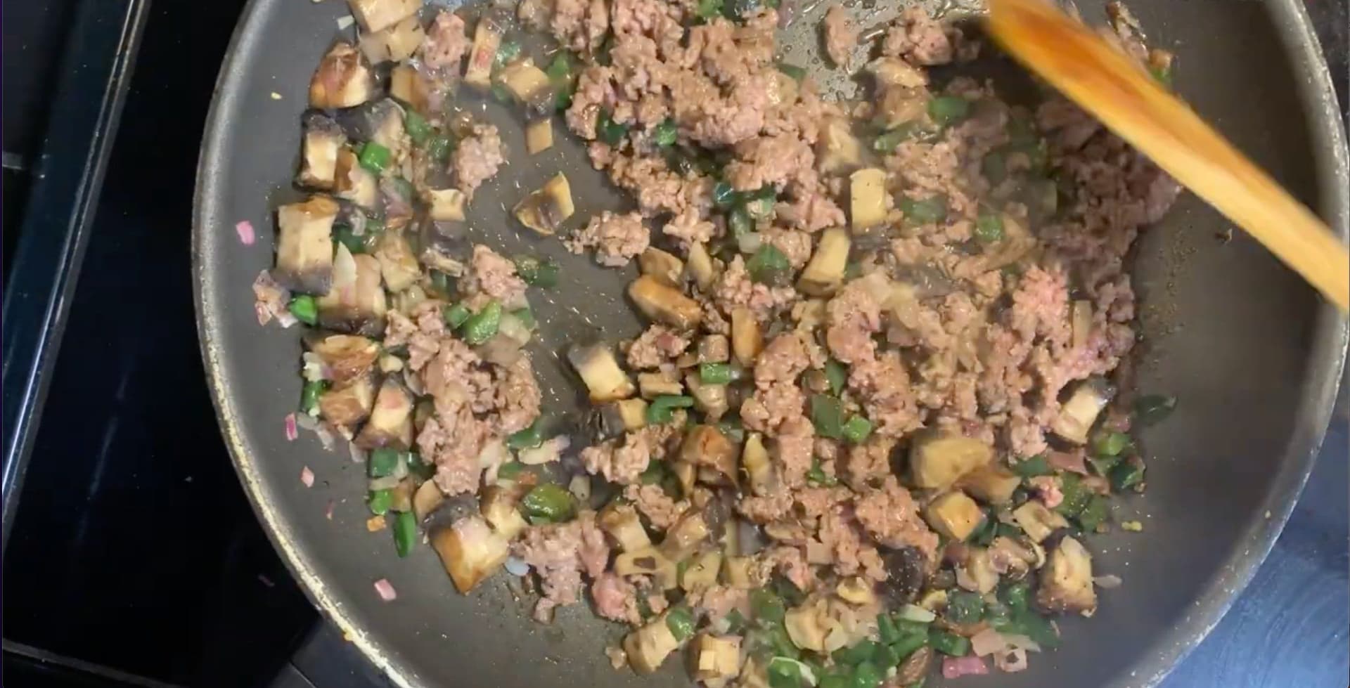Ground meat and diced mushrooms cooking in a pan with green onions, being stirred with a wooden spoon.
