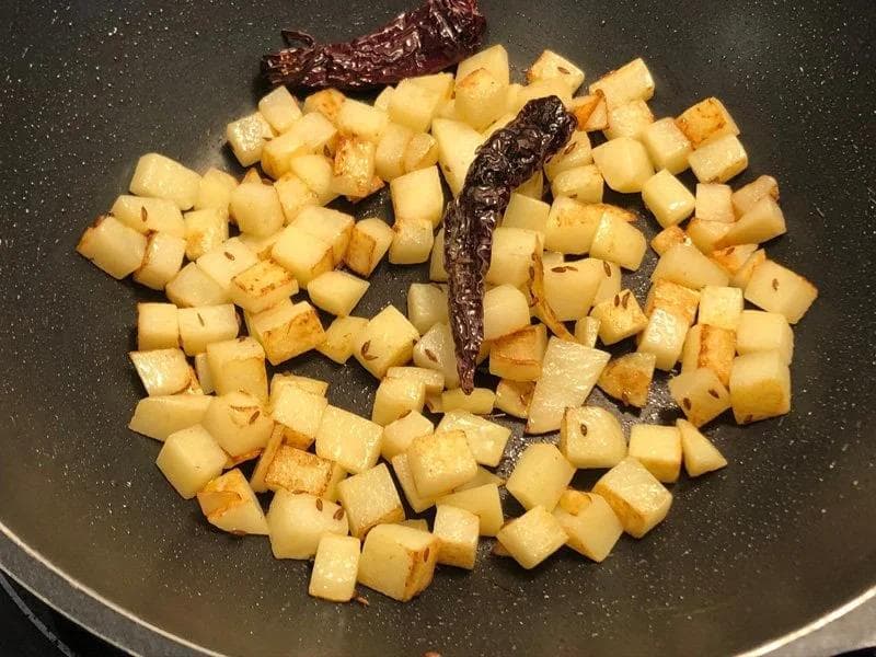Diced potatoes frying in a dark nonstick pan with cumin seeds and two dried red chilies.