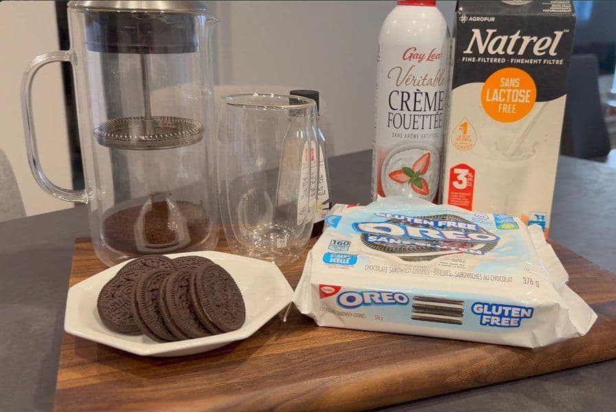 Ingredients for Oreo latte displayed on wooden board: Oreo cookies on white plate, milk carton, coffee creamer, and water pitcher with glasses.
