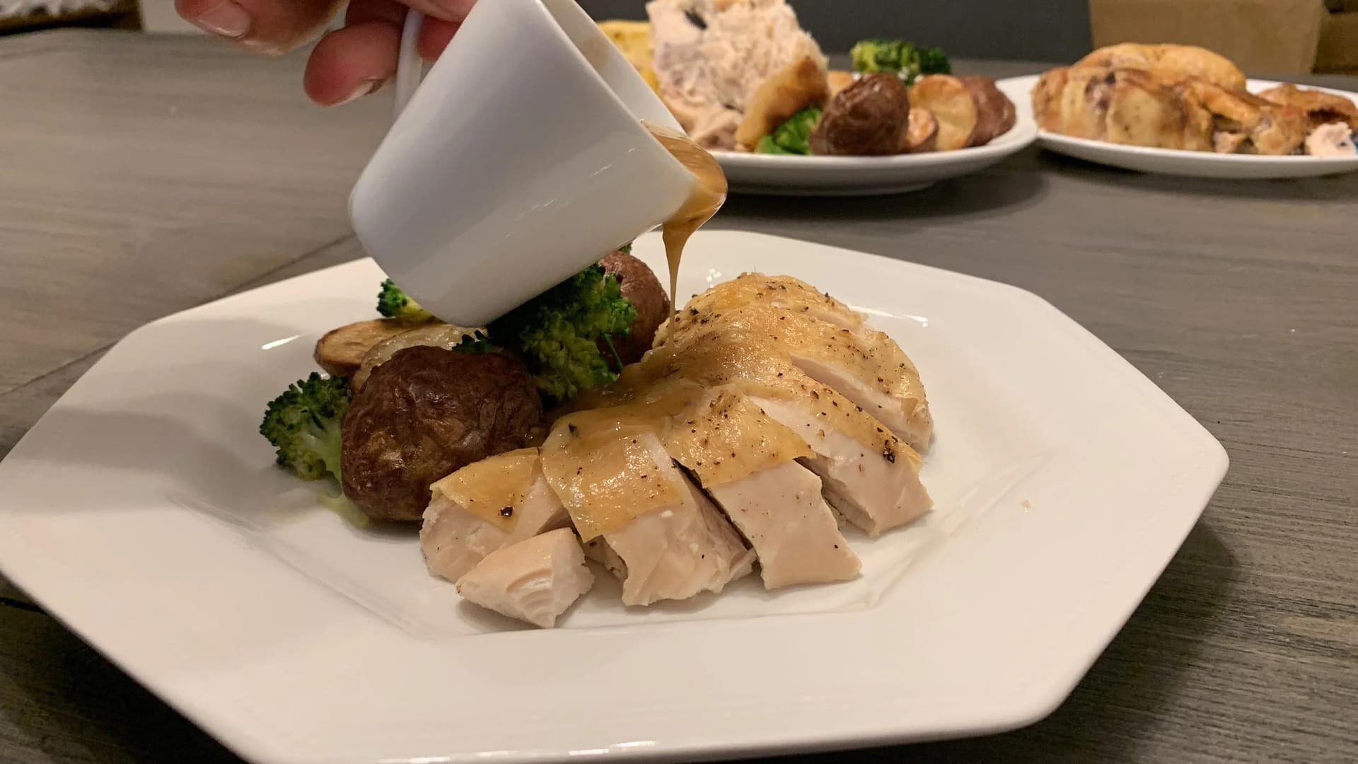 Sliced turkey breast with gravy being poured over it, served with roasted potatoes and broccoli on a white plate.