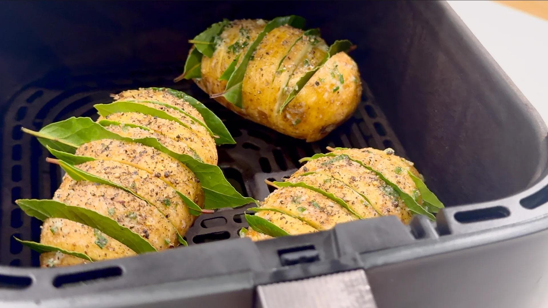Four hasselback potatoes in air fryer basket, sliced and stuffed with bay leaves, golden and partially cooked.