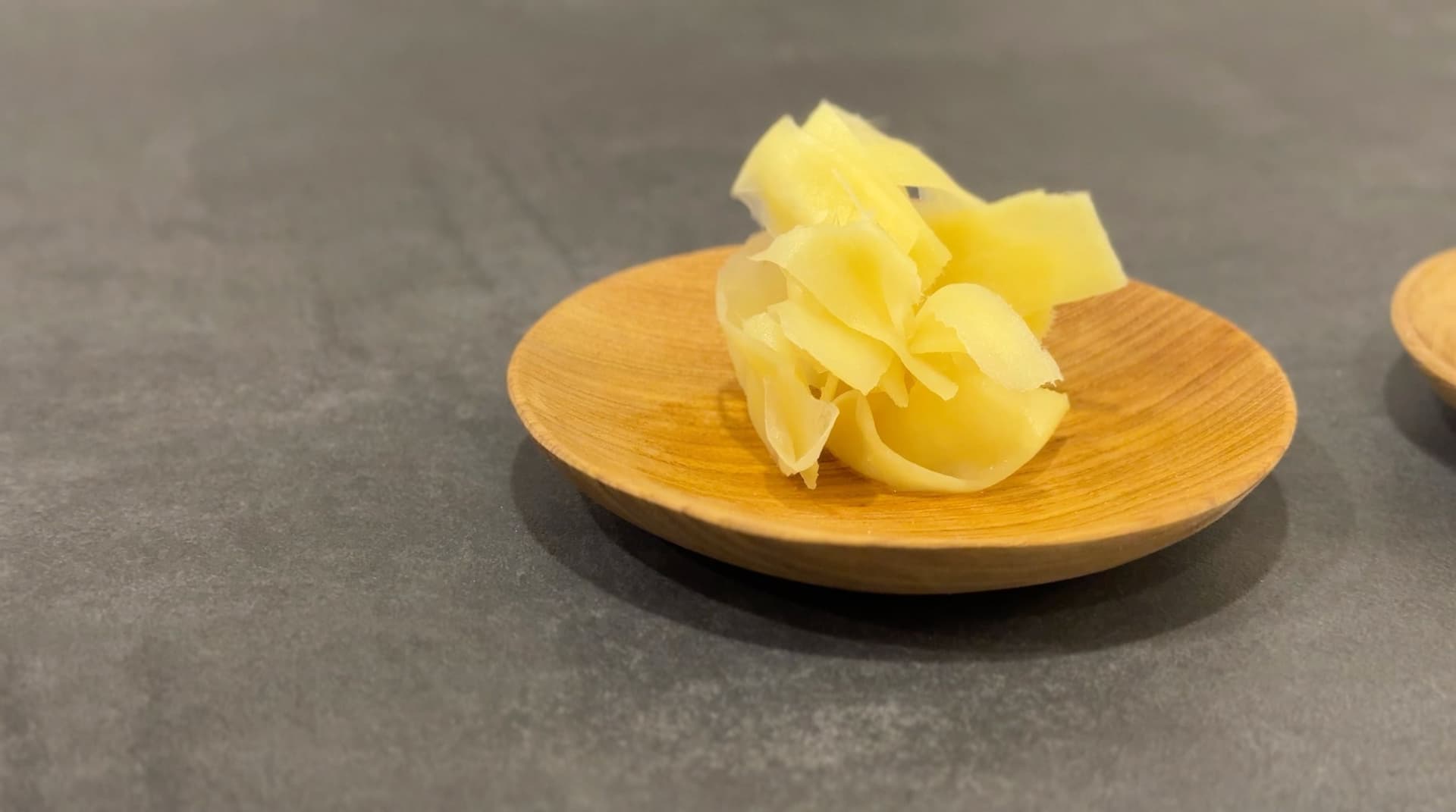 Pickled ginger slices arranged on a small wooden plate against a gray surface.