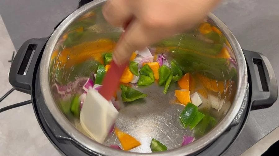 Hand adding red bell pepper to sautéed onions and peppers in Instant Pot on sauté mode.