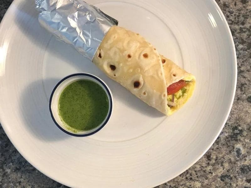 Spicy egg roll wrapped in foil served with green cilantro chutney on a white plate.
