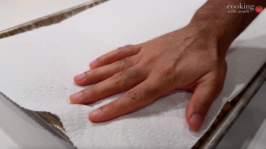 Hand patting dried chickpeas with paper towel on baking tray to remove excess moisture.