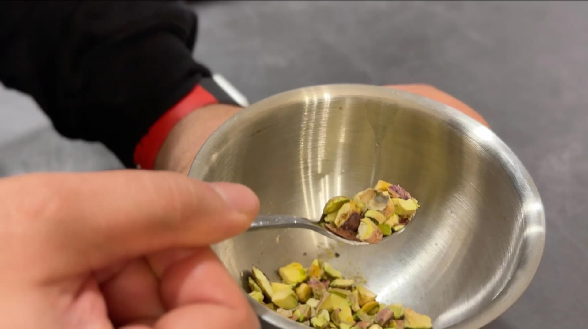 Spoon holding chopped pistachios over a metal bowl of creamy semolina pudding during preparation.