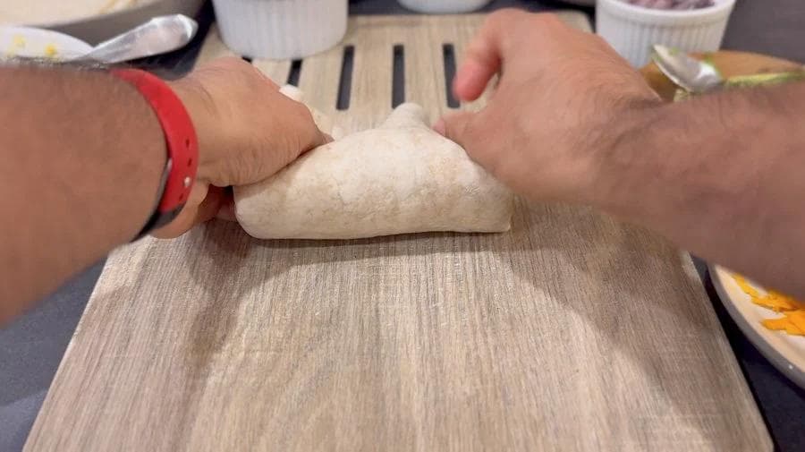 Hands kneading and folding a burrito filled with eggs, cheese, and sauce on a wooden surface.