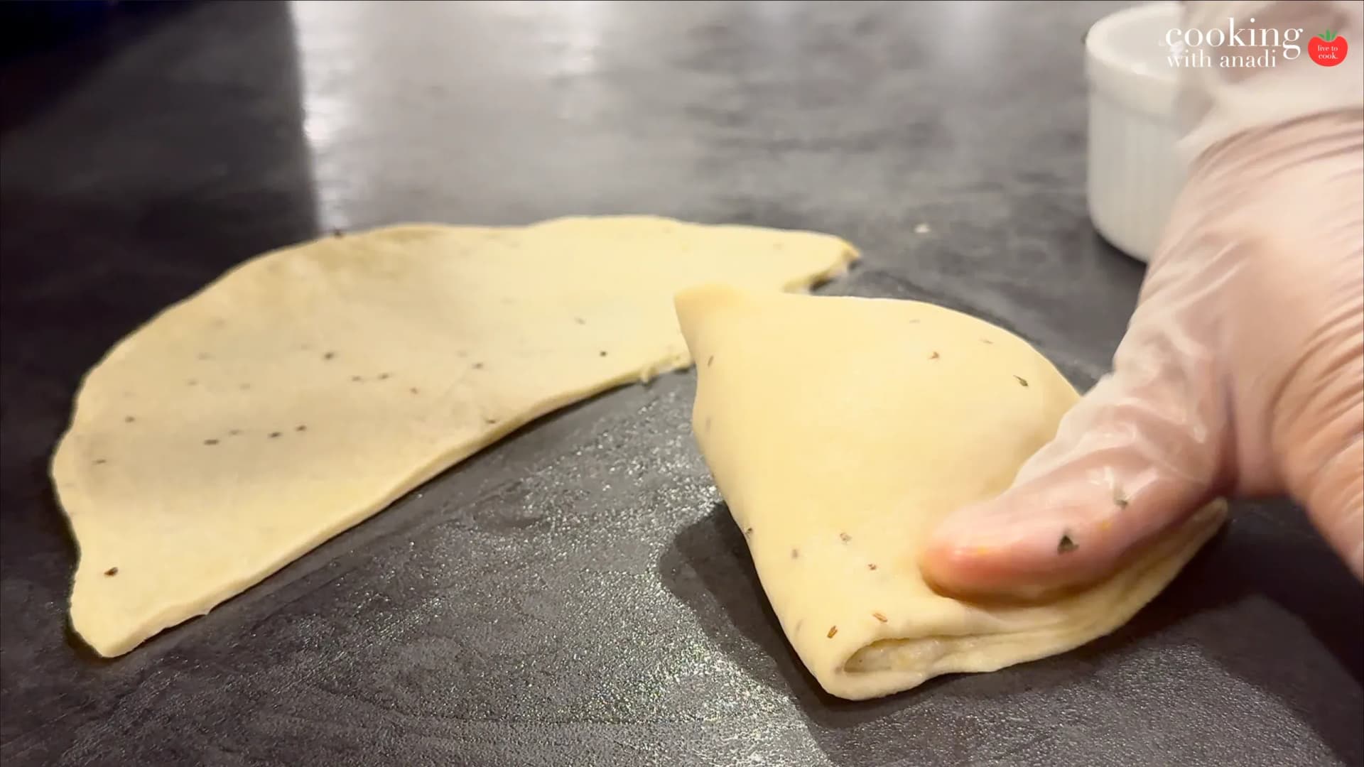 Hands folding and sealing potato samosa dough into triangular shapes on a dark surface.