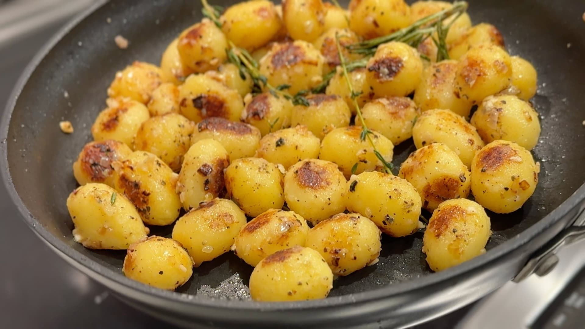 Golden sautéed baby potatoes with rosemary and herbs in a dark skillet, showing caramelized edges and browning.