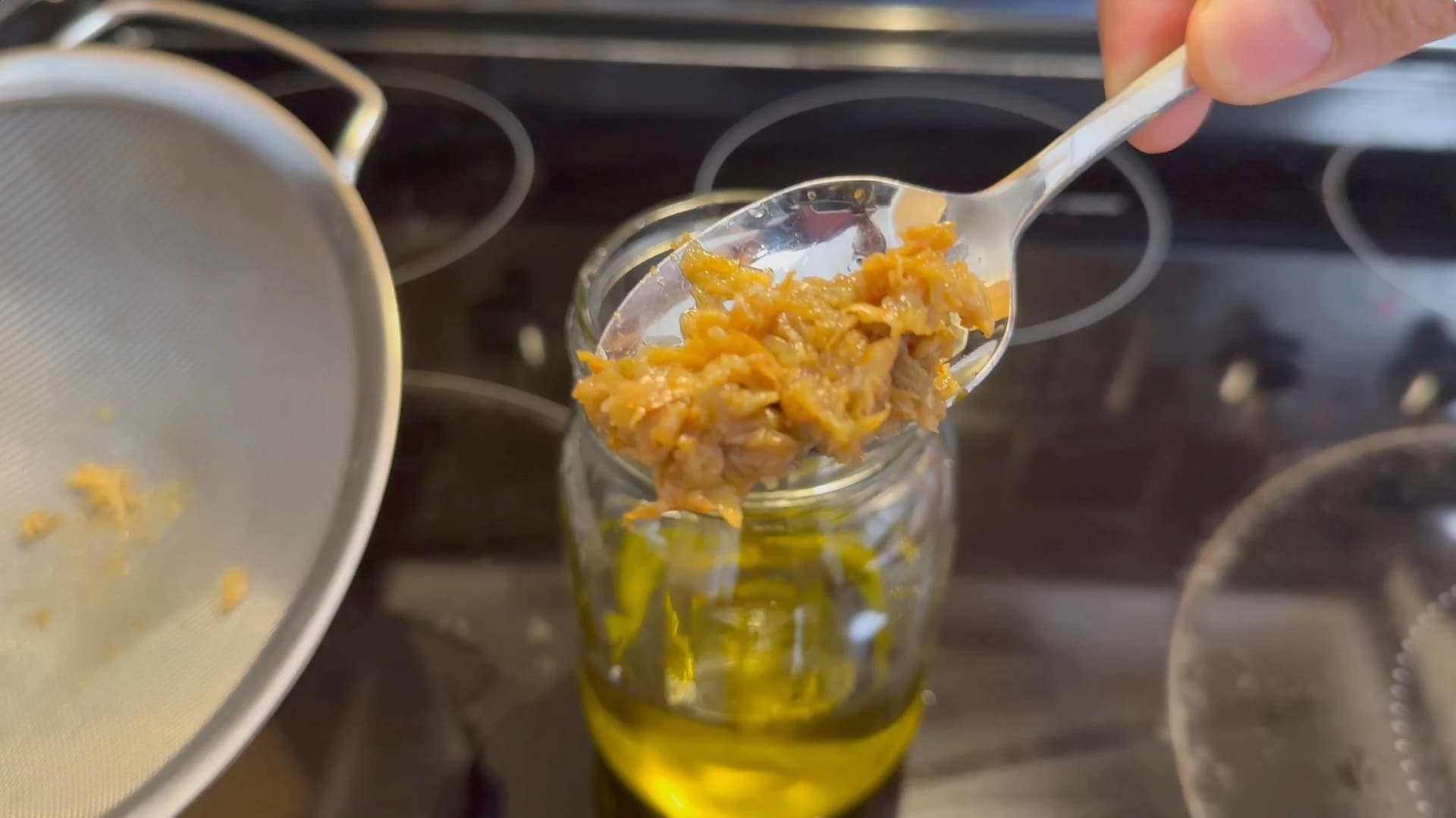 Spoon lifting golden roasted garlic pieces from olive oil in a glass jar during preparation of garlic-infused oil.