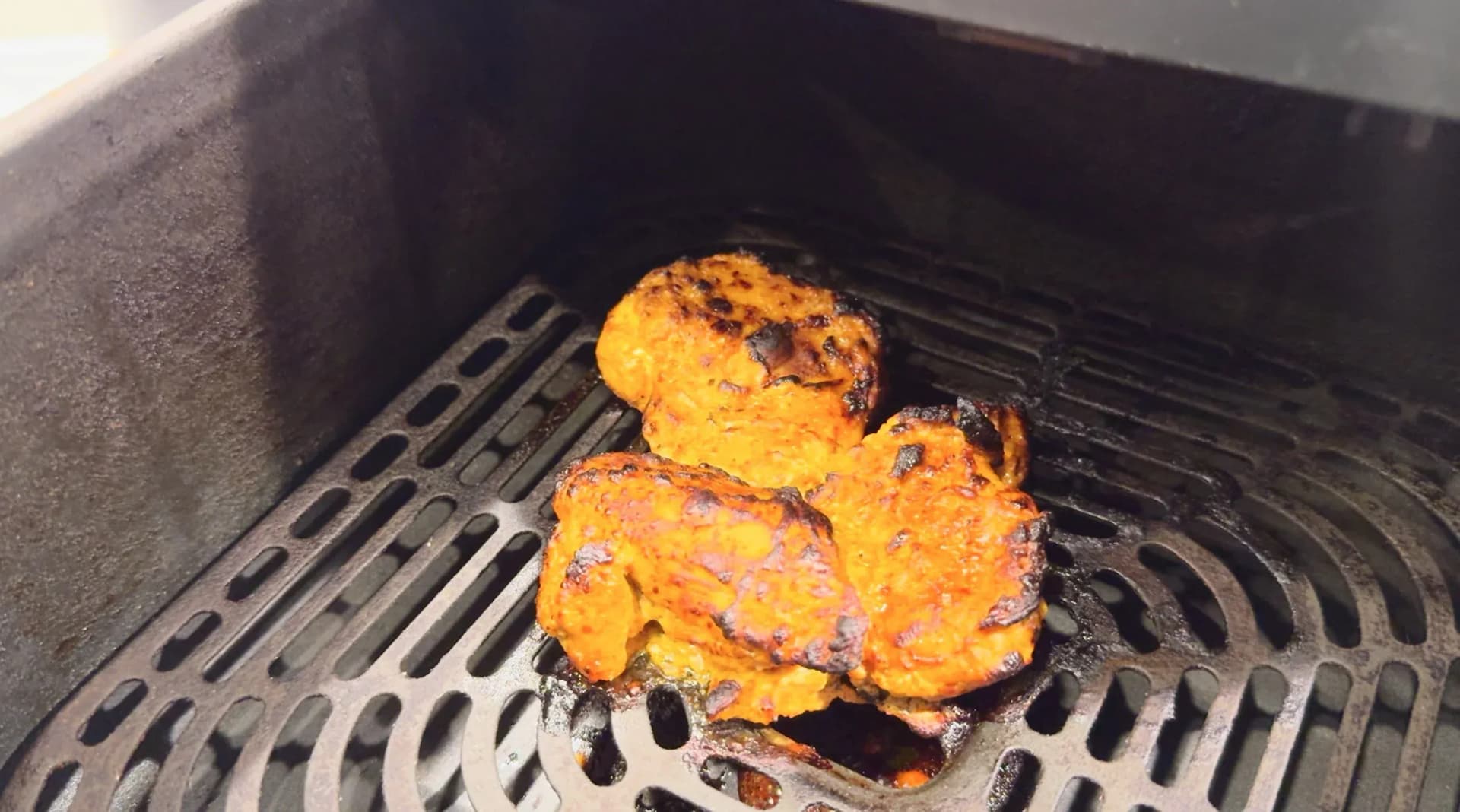 Overcrowded air fryer basket with touching pieces of marinated chicken tikka showing charred orange-spiced coating.