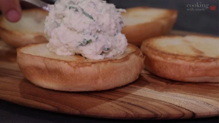 Crab salad being spread onto toasted buns on a wooden serving board for sandwiches.
