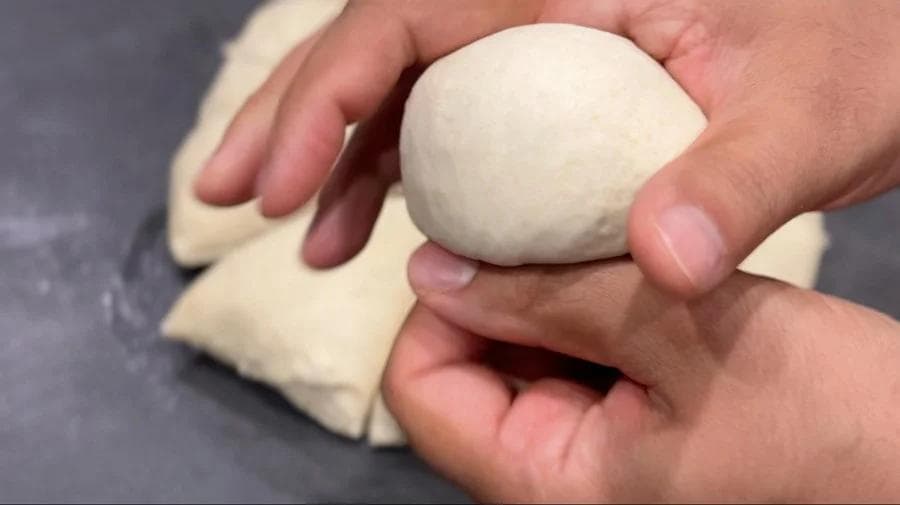 Hands shaping a ball of pita dough, with another flattened portion visible on the work surface.