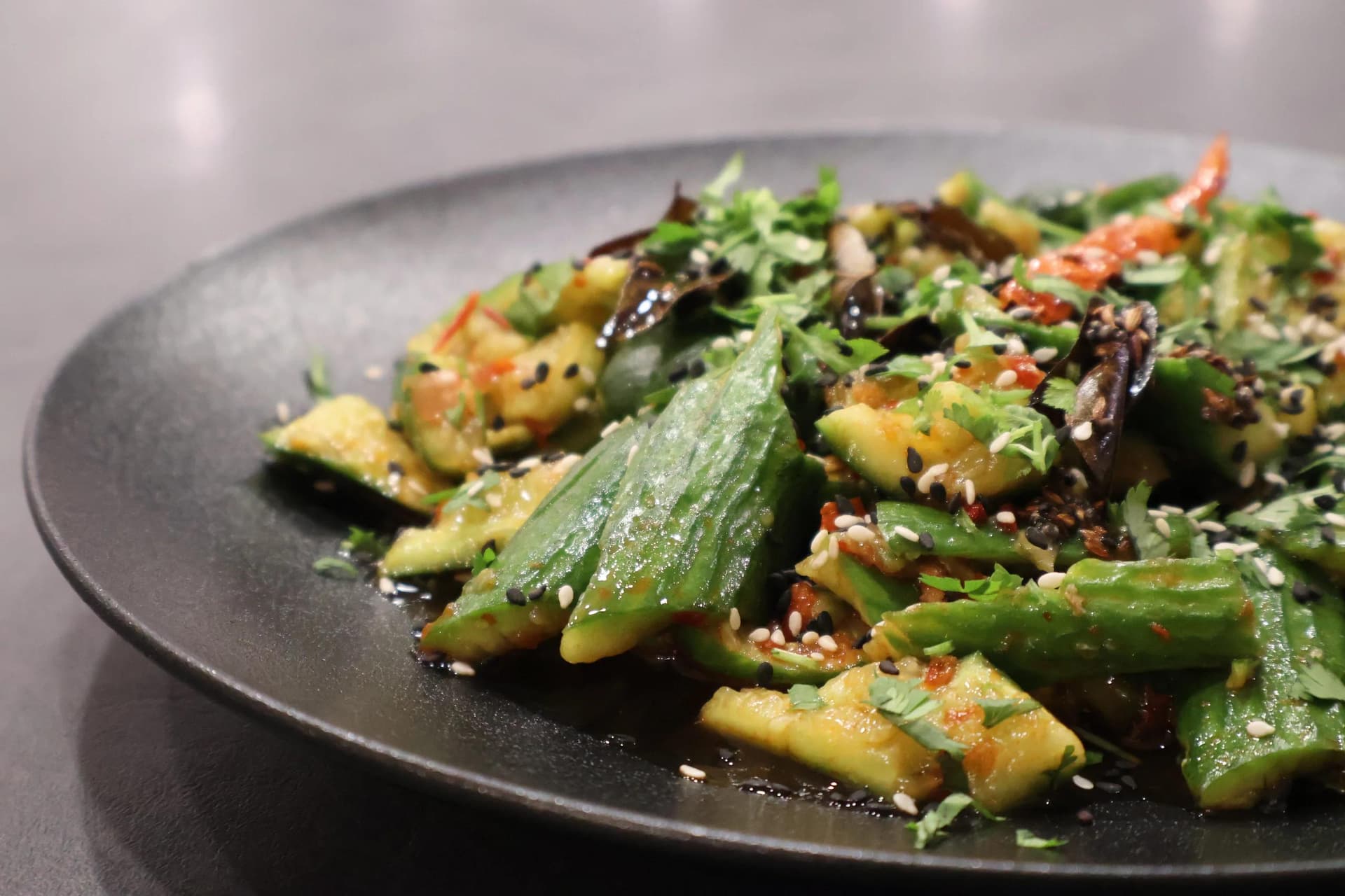 Indian fusion smashed cucumber salad with sesame seeds, red chili, and fresh herbs on a dark plate.