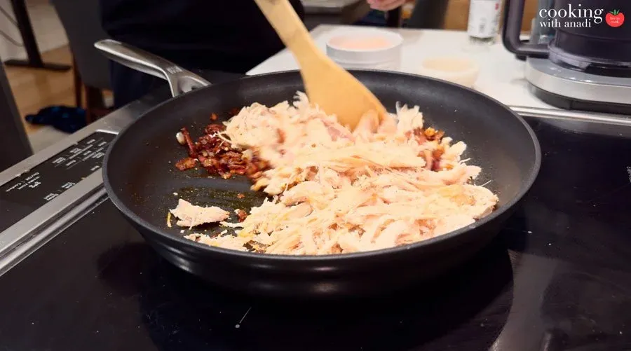 Shredded chicken and sautéed spiced onions being stirred together in a black nonstick skillet with a wooden spatula.