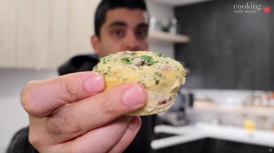 Hand holding a baked egg muffin with herbs and vegetables visible on top.