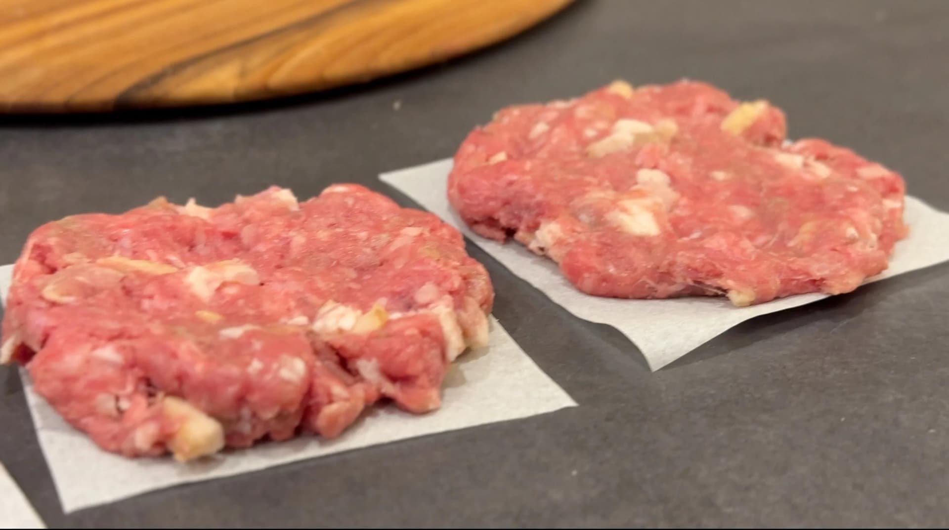 Two raw burger patties on parchment paper, topped with bacon and cheese pieces, ready for cooking.