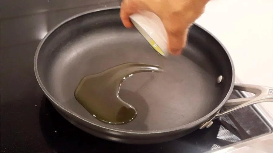 Olive oil being poured into a dark skillet on a stovetop for sautéing onions and garlic.