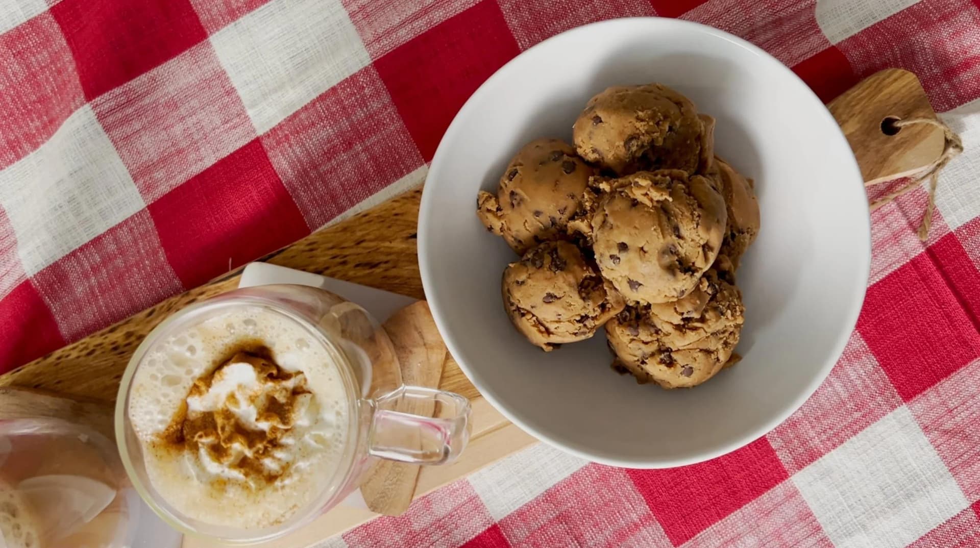 Chocolate chip edible cookie dough served in a white bowl with a mug of whipped dough topped with cinnamon on a red gingham cloth.