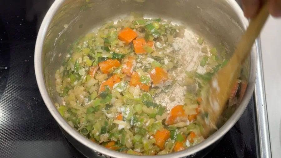 Sautéed vegetables including diced onions, carrots, leeks, and garlic in a white pot with flour coating.