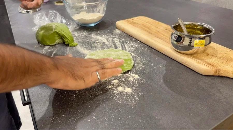 Hand folding a green spinach dough portion dusted with flour on a dark countertop during palak paratha preparation.