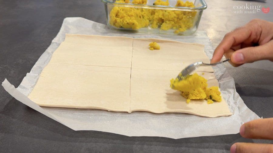 Brushing egg wash onto a potato-filled puff pastry turnover with a pastry brush on parchment paper.