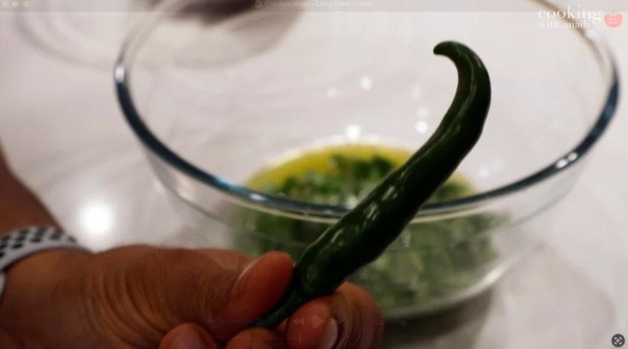 A hand holding a fresh green chili pepper over a glass bowl containing green marinade ingredients.