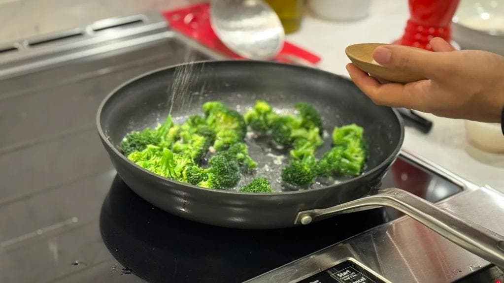 Broccoli florets cooking in a black pan with garlic butter, being stirred with a wooden spoon.