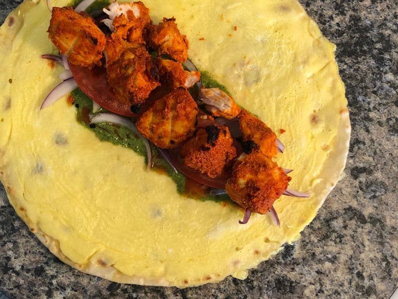 Chicken tikka pieces with purple onions and cilantro on a yellow paratha bread on a gray surface.