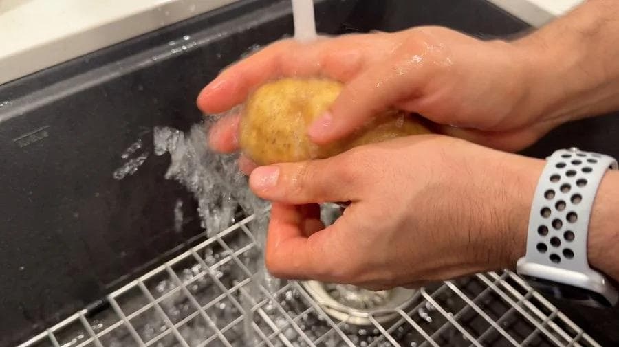 Hands washing a golden potato under running water in a kitchen sink.