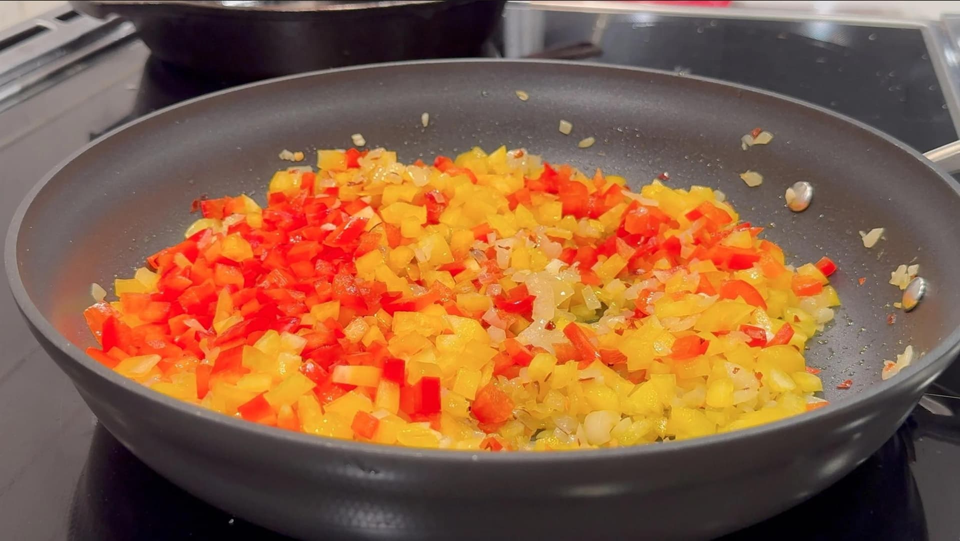 Diced red and yellow bell peppers sautéing with onions in a large non-stick skillet on a stovetop.