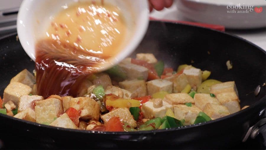 Glossy sauce being poured over cubed tofu and vegetables in a wok during stir fry preparation.