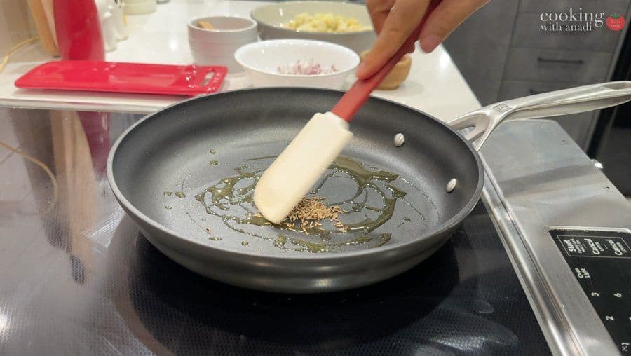 Tempering spices in hot oil in a skillet with a spatula, showing cumin seeds and aromatics being sautéed.